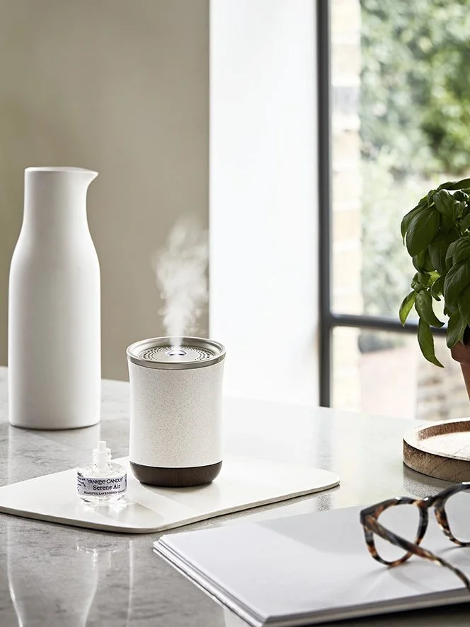 A white essential oil diffuser emitting mist on a white tray, next to a small bottle of essential oil, with glasses, a notebook, and a plant nearby, near a window.