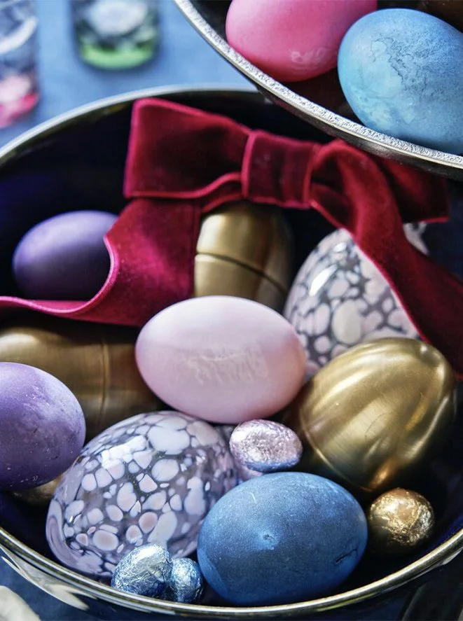 A collection of decorated Easter eggs in a bowl, with some resting on a red velvet cloth. The eggs are in various pastel and metallic colors, including pink, blue, purple, and gold, some with speckled or marbled patterns.