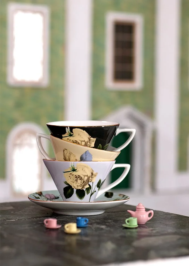 Three stacked decorative teacups with floral patterns on a saucer, surrounded by miniature teapots and cups on a wooden surface with a blurred green building in the background.