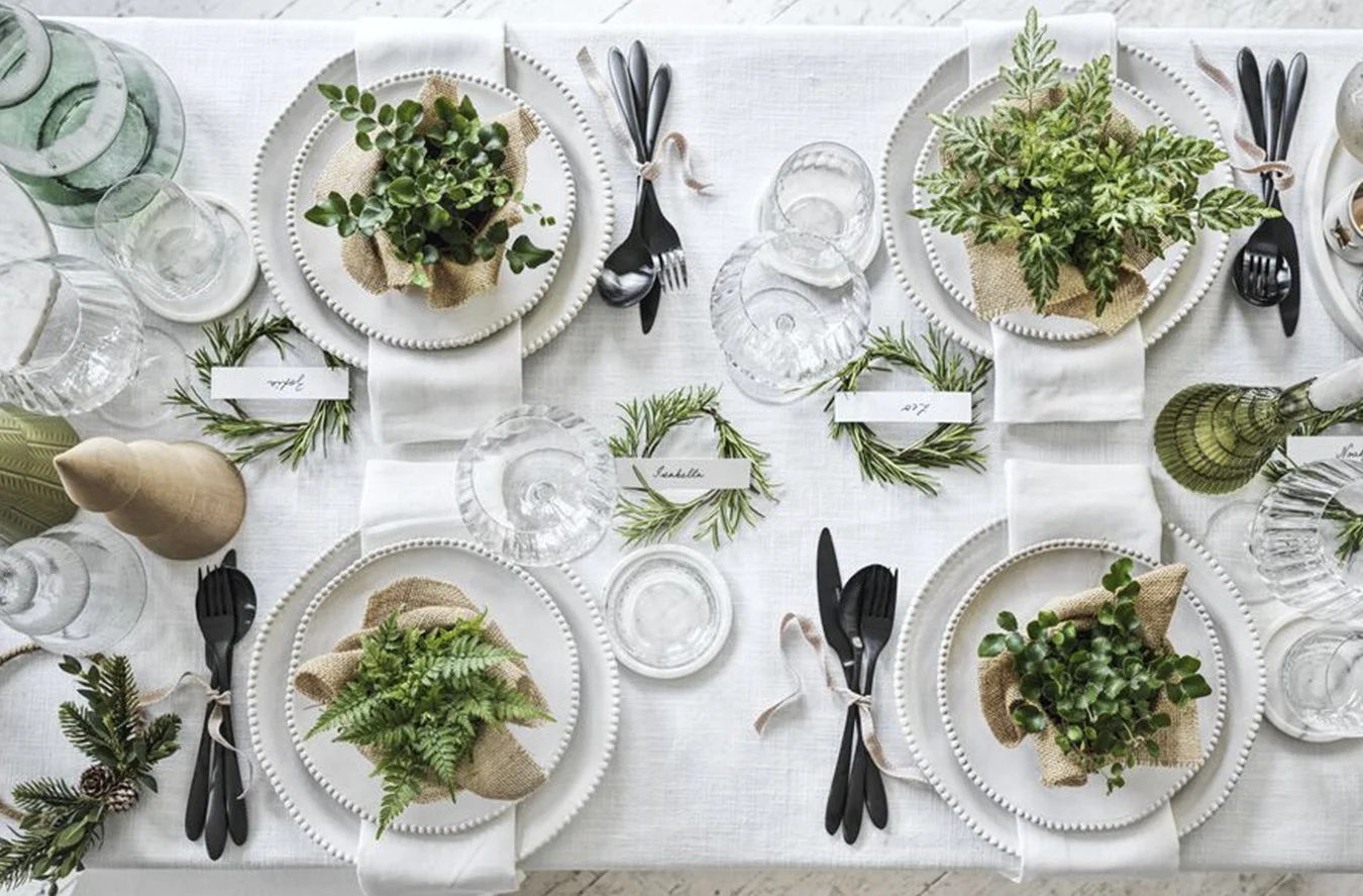 A festive table setting with white plates, black cutlery tied with ribbons, glassware, and greenery centerpieces on a white tablecloth.