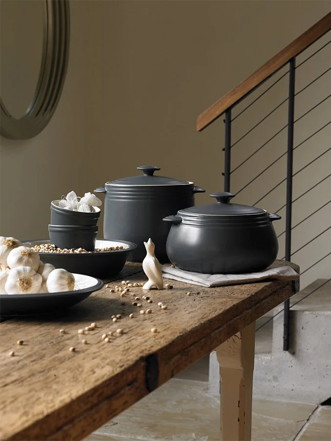 A rustic wooden table with black ceramic pots, some white garlic bulbs, a bowl of beans or grains, and a small ceramic rabbit figurine, set in a home interior with a staircase railing in the background.