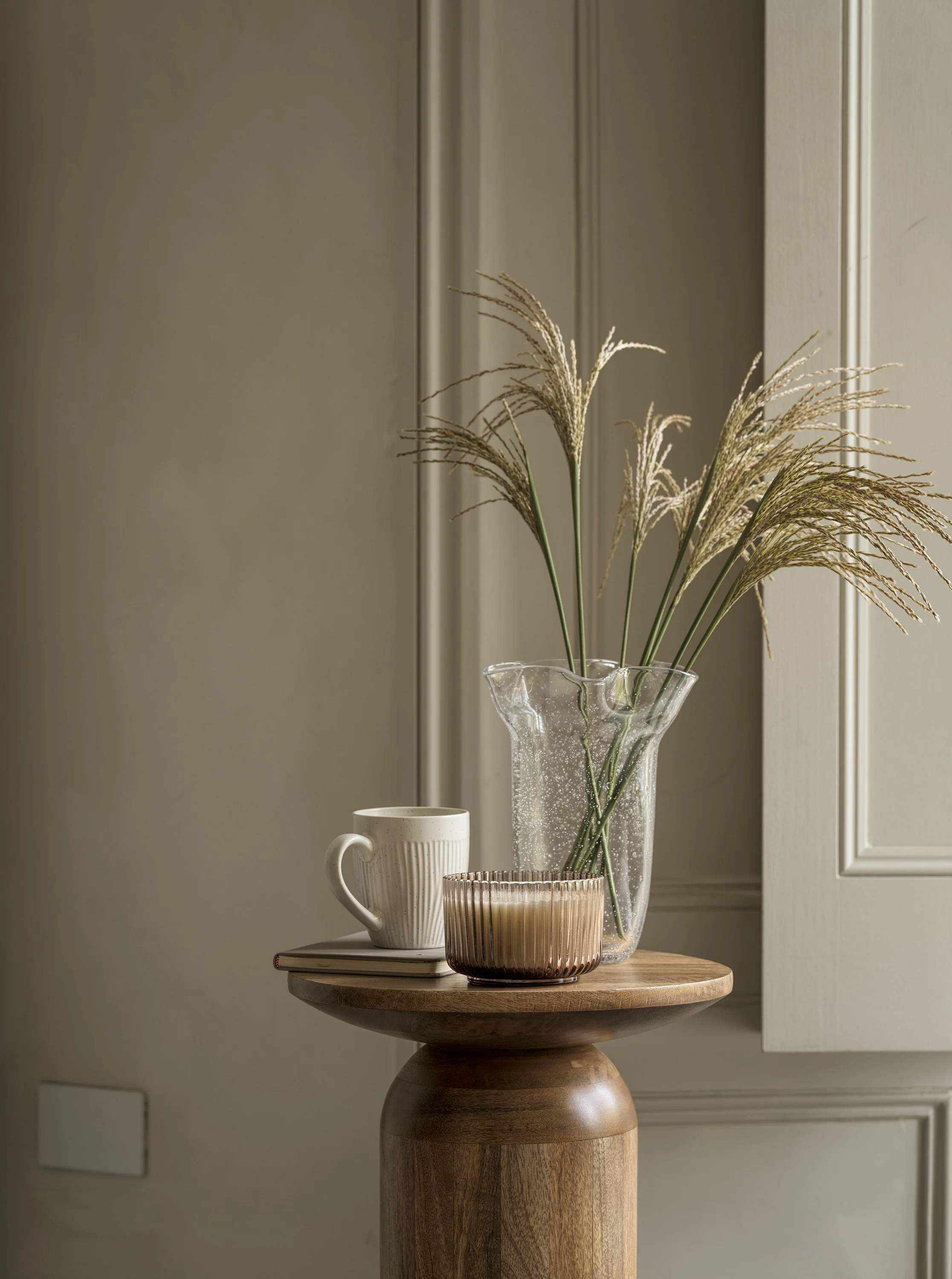 Decorative arrangement on a wooden round table with a tall glass vase holding dried grasses, a textured beige candle holder, and a white mug, against a neutral wall.