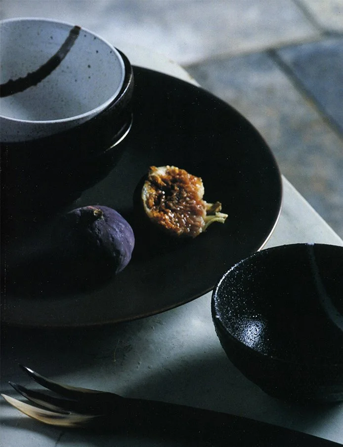 A black plate with a cut-open fig and a whole purple fig, a white teacup with a dark liquid, a black bowl with a dark liquid, and a silver fork on a gray marble surface with natural light.