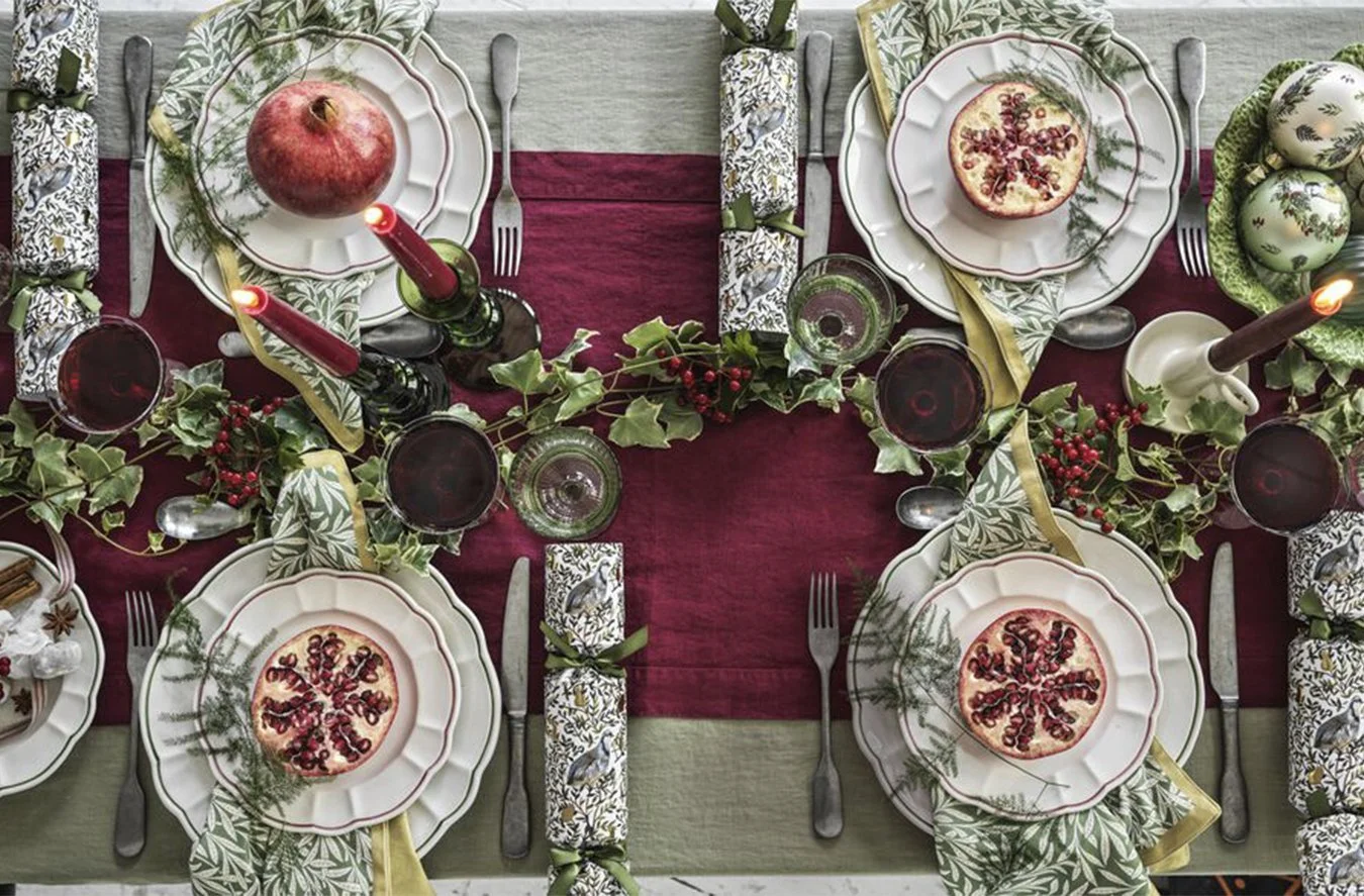 A festive holiday table setting with plates topped with pomegranate and raspberry decorations, green and red elements, and candles, indicating a Christmas or holiday celebration.
