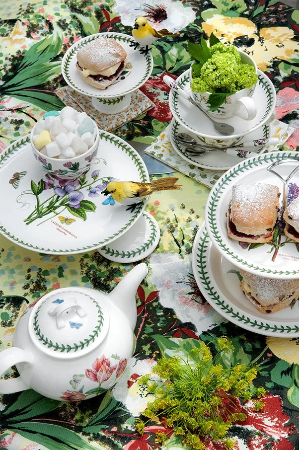 A table set with teacups, saucers, plates, and a teapot, featuring a floral design with green leaves and purple flowers. On the table are cupcakes with powdered sugar, small bowls of marshmallows, a small yellow bird figurine, and a green plant in a 
