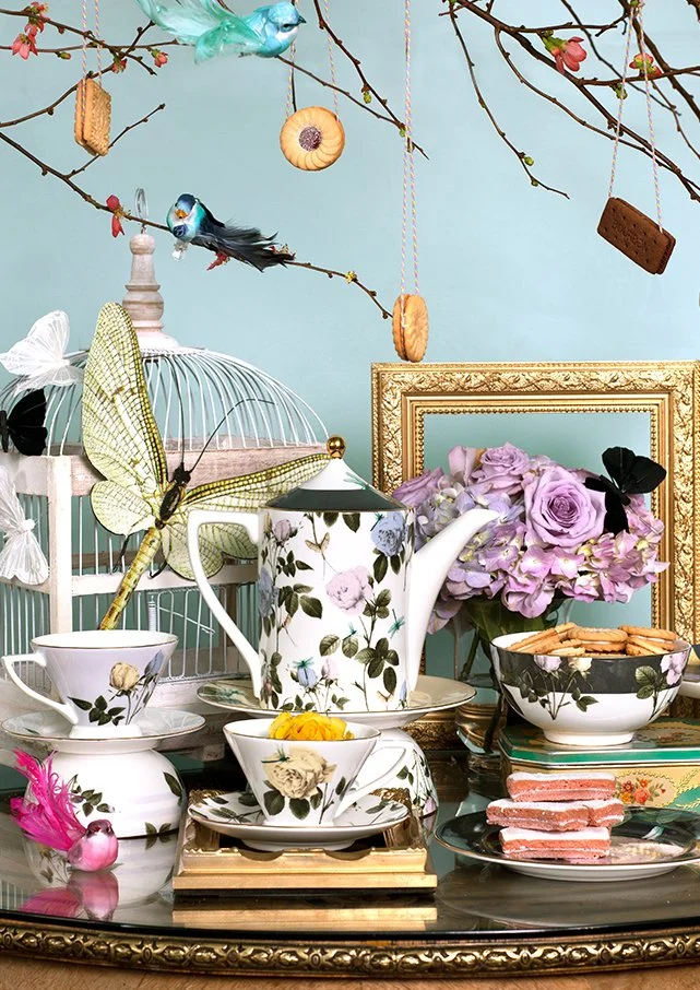 Decorative tea set with floral patterns, a birdcage with a black-and-blue bird, pink flowers, colorful hanging bird ornaments, cupcakes, and snacks on a black table in front of a blue wall with an empty gold picture frame.