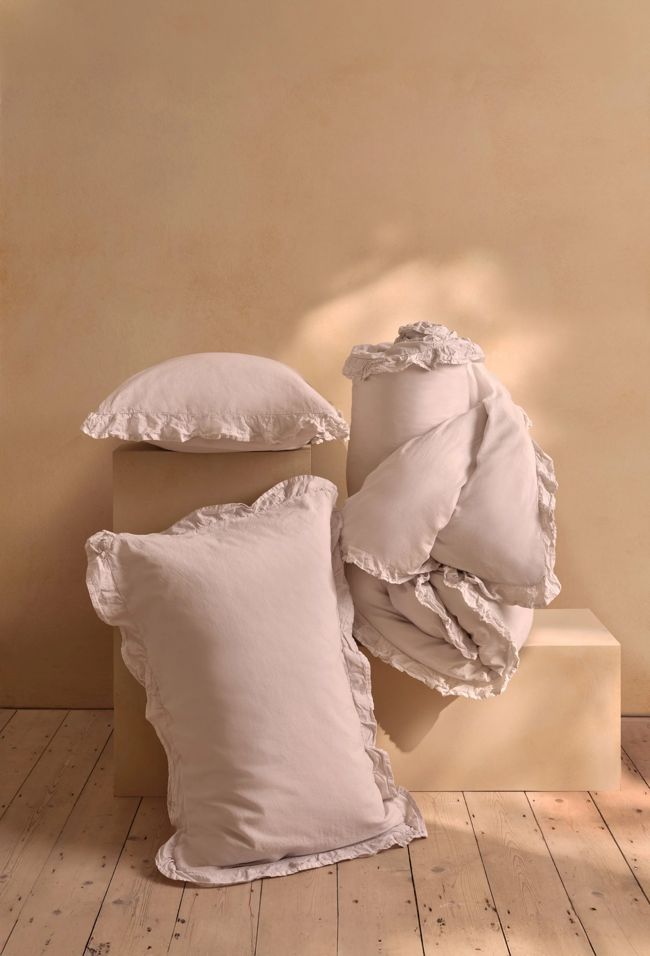 A collection of beige pillows with ruffled edges, arranged on and around a beige platform against a plain beige wall and wooden floor.