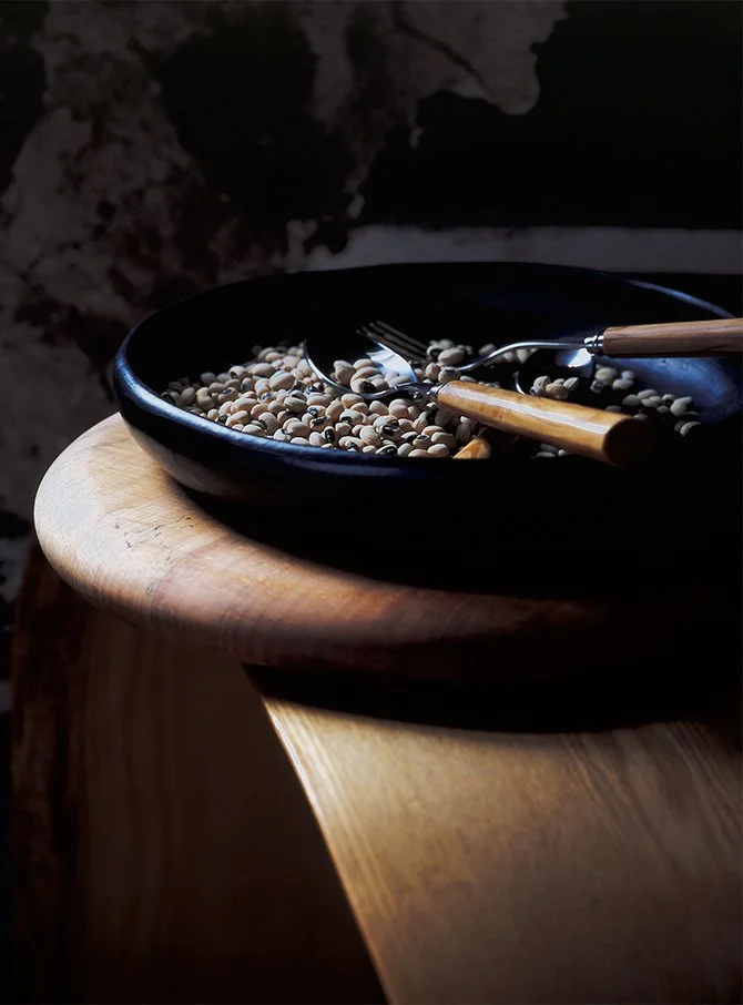 A black bowl containing white and black peppercorns on a wooden table, with a fork and spoon resting inside, in a dimly lit setting.