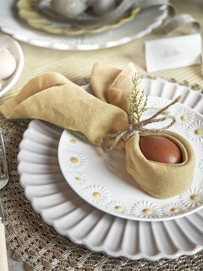 A white decorative plate with a floral pattern holds a cloth napkin wrapped around a brown egg, tied with twine and adorned with small sprigs of greenery, set on a table with a lace tablecloth.