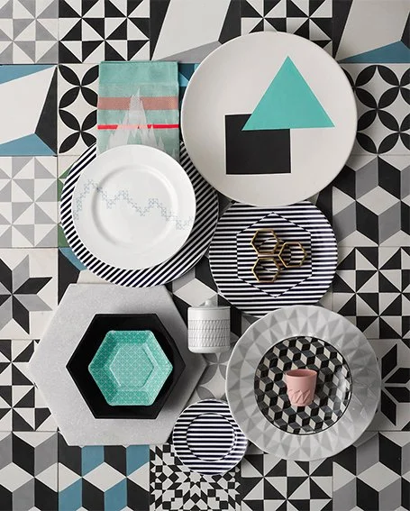 A collection of decorative plates, bowls, and tableware arranged on a black and white geometric patterned surface.