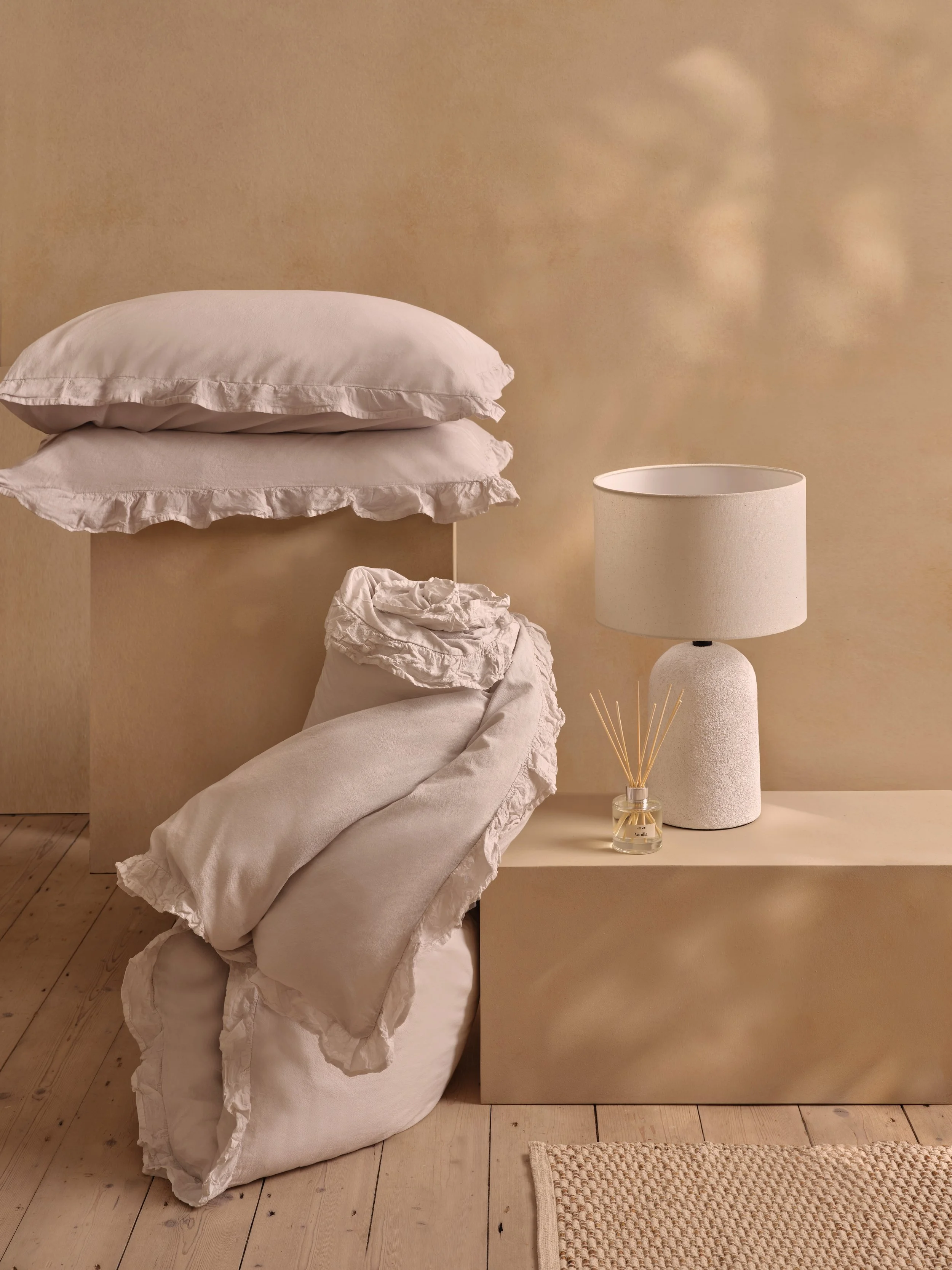 A cozy bedroom corner with a pile of beige pillows and a beige blanket on the floor next to a modern beige bedside table. On the table, there is a white textured table lamp and a small glass jar with reed diffuser sticks, set against a warm, neutral-