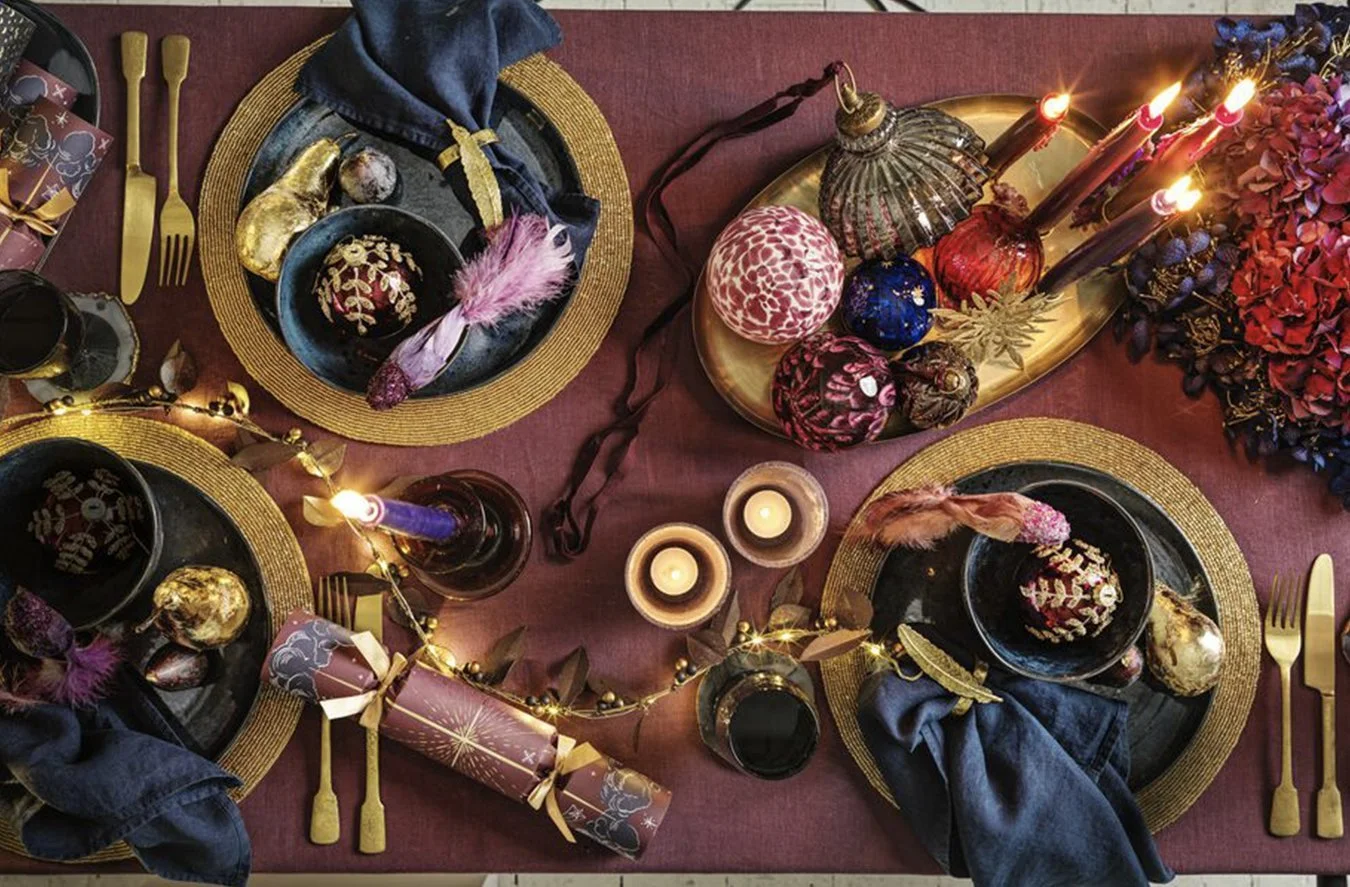 A festive holiday table setting with gold and black plates, blue cloth napkins, gold cutlery, lit candles, Christmas ornaments, and decorative elements arranged on a maroon tablecloth.