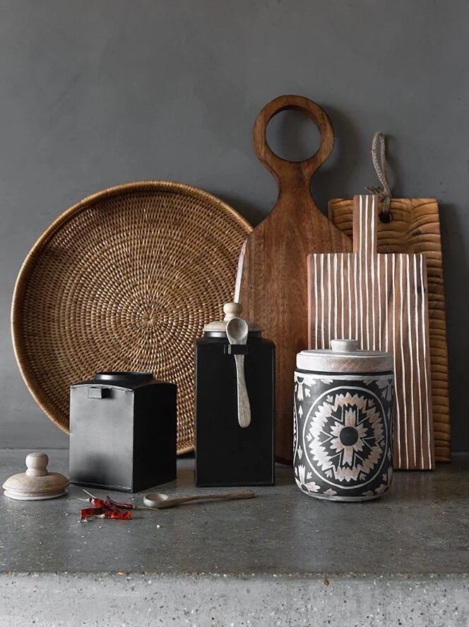 Kitchen countertop with wicker tray, wooden cutting boards, black containers, ceramic canister, spoon, and small decorative items.