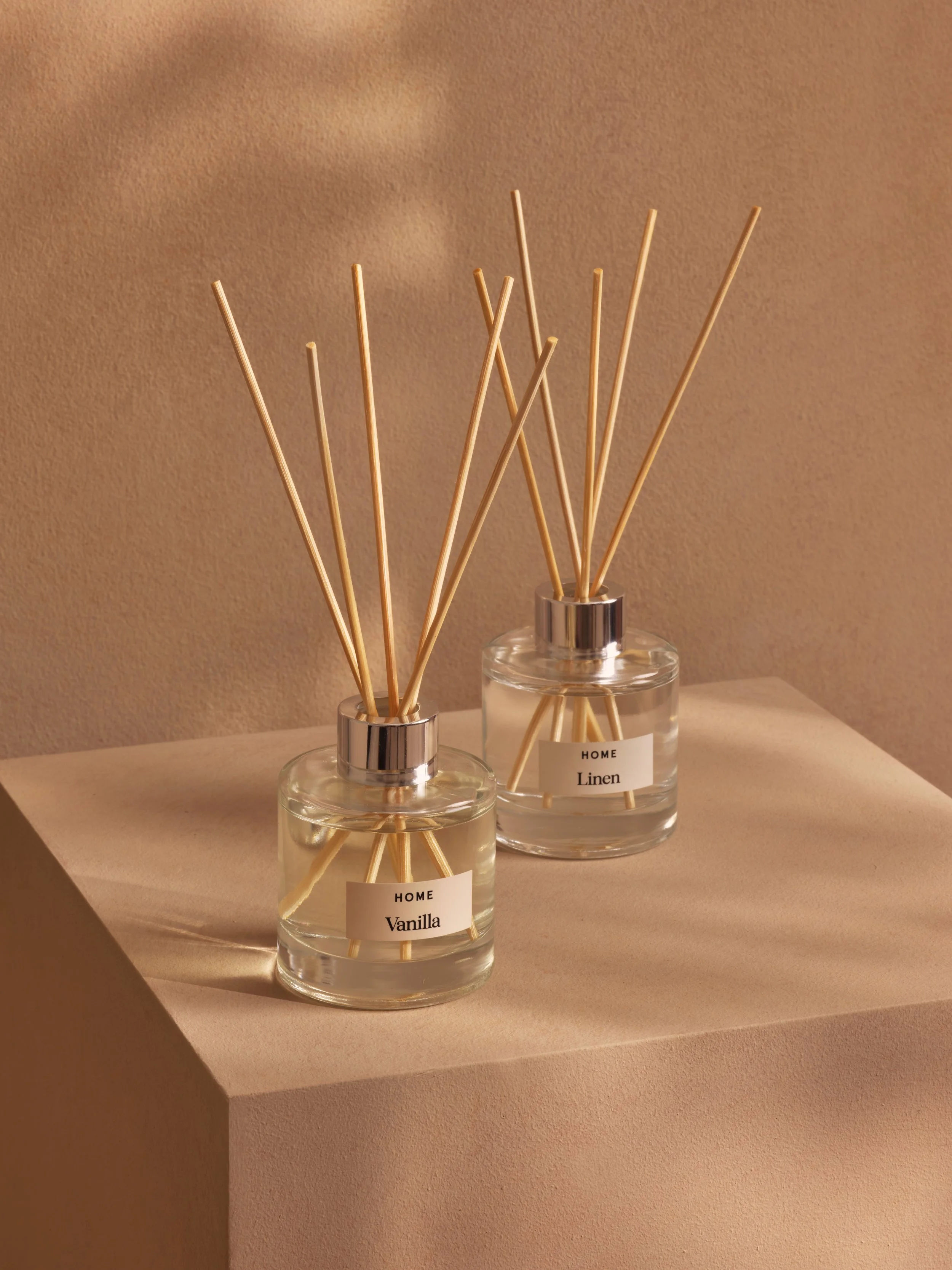 Two glass reed diffusers labeled 'Vanilla' and 'Linen' on a beige surface, with wooden sticks inside.