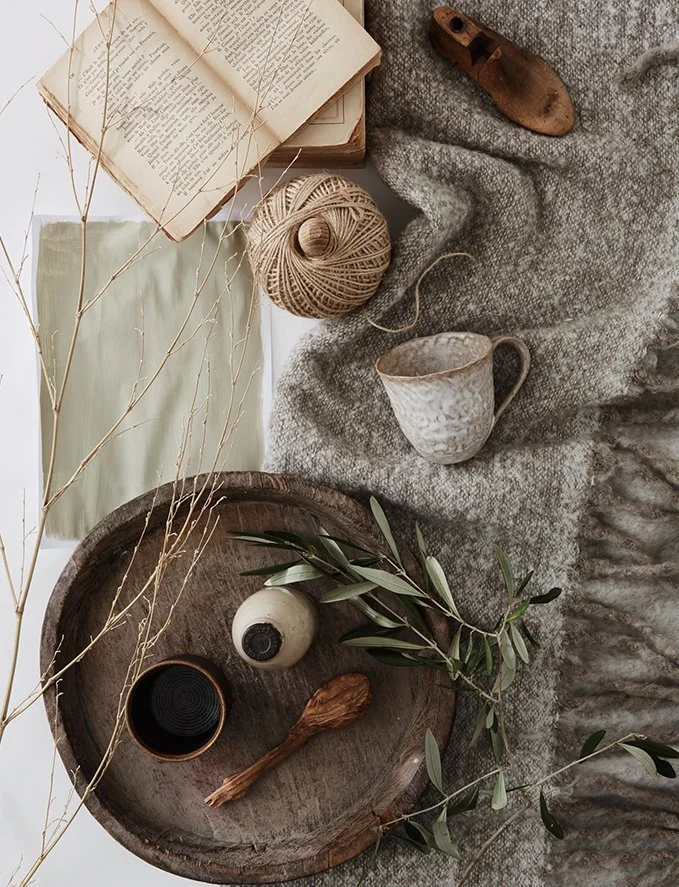 Rustic flat lay of vintage books, a ceramic mug, a ball of twine, dried branch, a wooden spoon, a glass deerskin, and a cloth on a textured surface with green leaves.