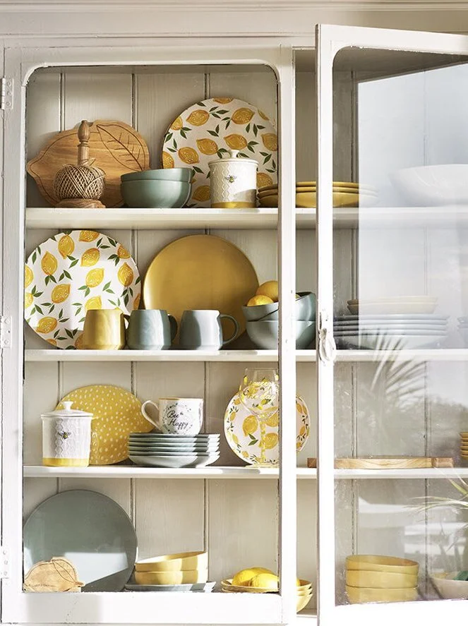Display cabinet filled with yellow, white, green, and lemon-themed dishware, including plates, cups, bowls, and pitchers.