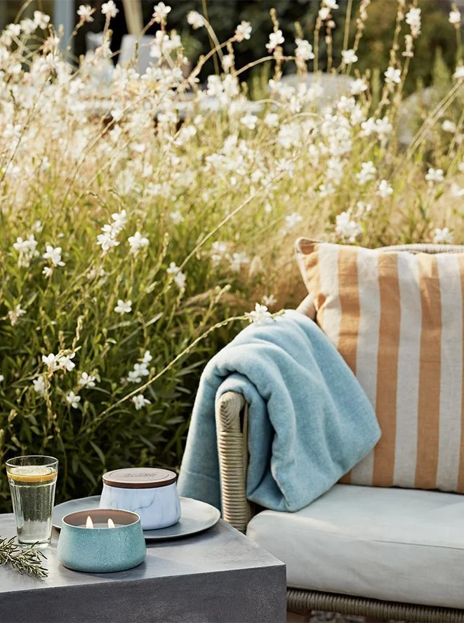 An outdoor scene with a cushioned wicker chair, a light blue blanket, a striped pillow, and a small table with a candle, a glass of water with lemon, and a decorative jar, set against a background of blooming white flowers and greenery.