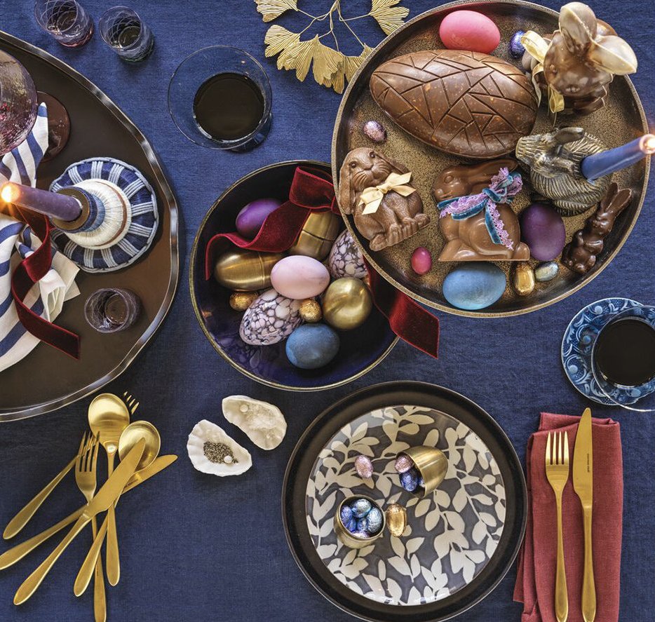 Easter table setting with decorated chocolate eggs, candy rabbits, and blue, pink, purple, and gold-colored candies on black and gold trays. Gold utensils and glasses of dark beverages on a dark fabric tablecloth with some decorative leaves.