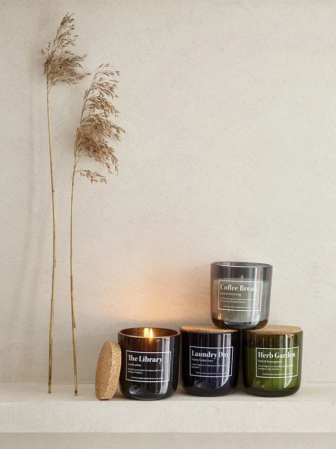 Decorative candles with labels reading "The Library," "Laundry Day," "Coffee Break," and "Herb Garden" arranged on a shelf with a rock, dried pampas grass, and wall in the background.