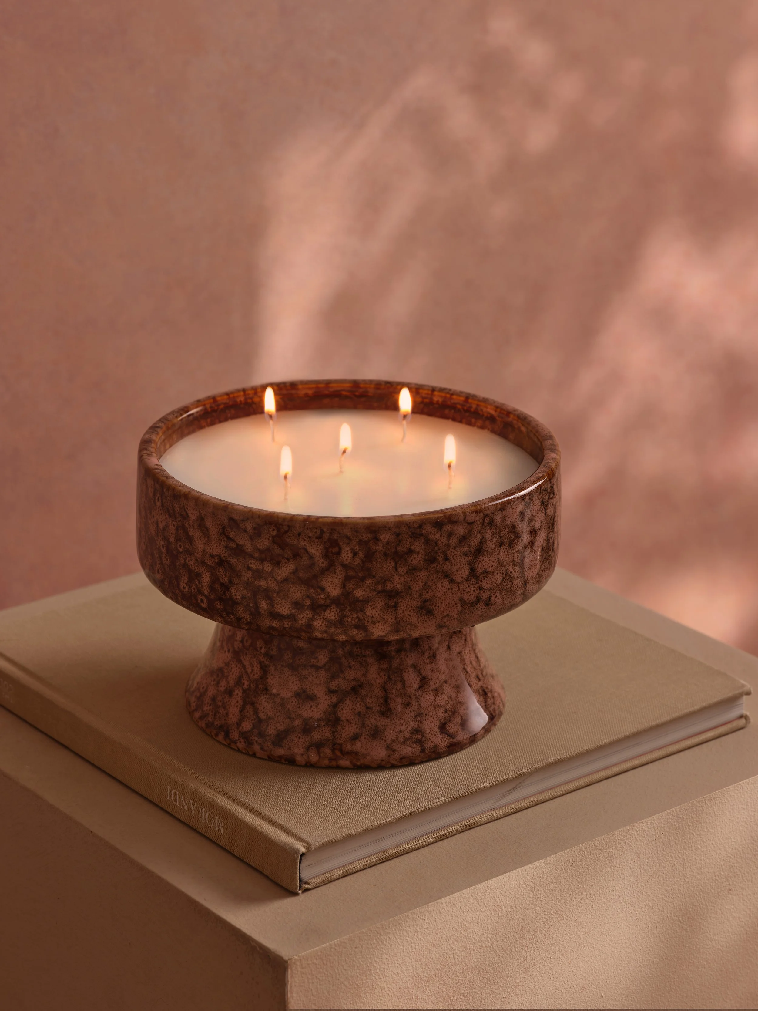 A stone candle holder with five lit wicks on a closed book, placed on a beige surface with a soft, pinkish background.