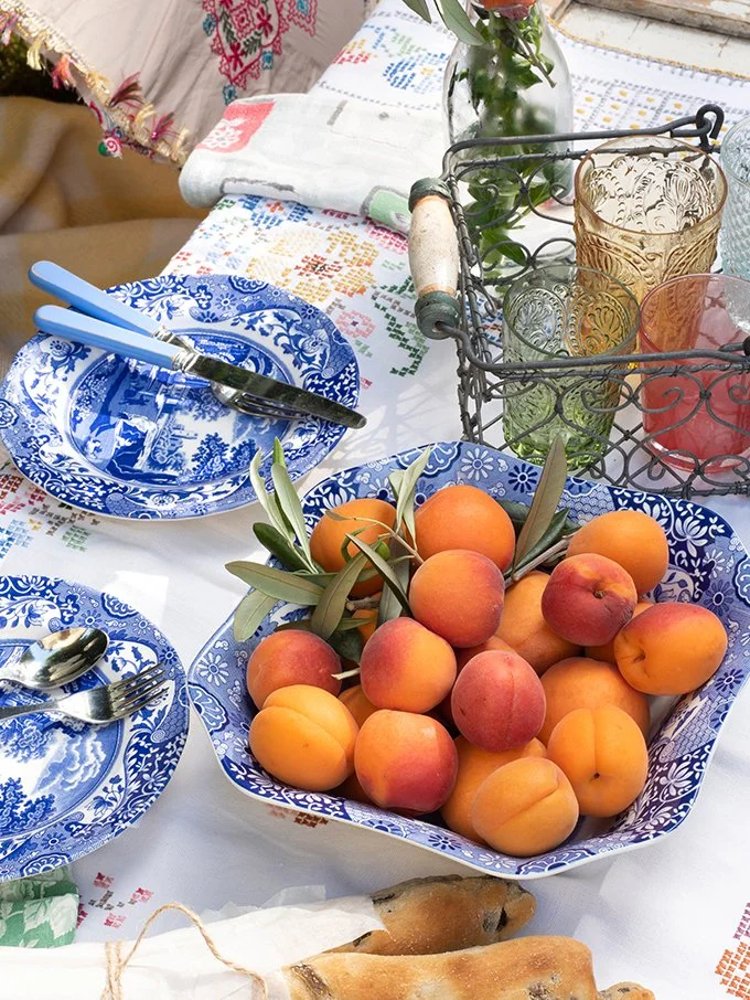 A table set for a meal with a large bowl of peaches, blue and white patterned plates, silverware, colorful glasses, and a vase with flowers. The tablecloth has a traditional embroidered design.