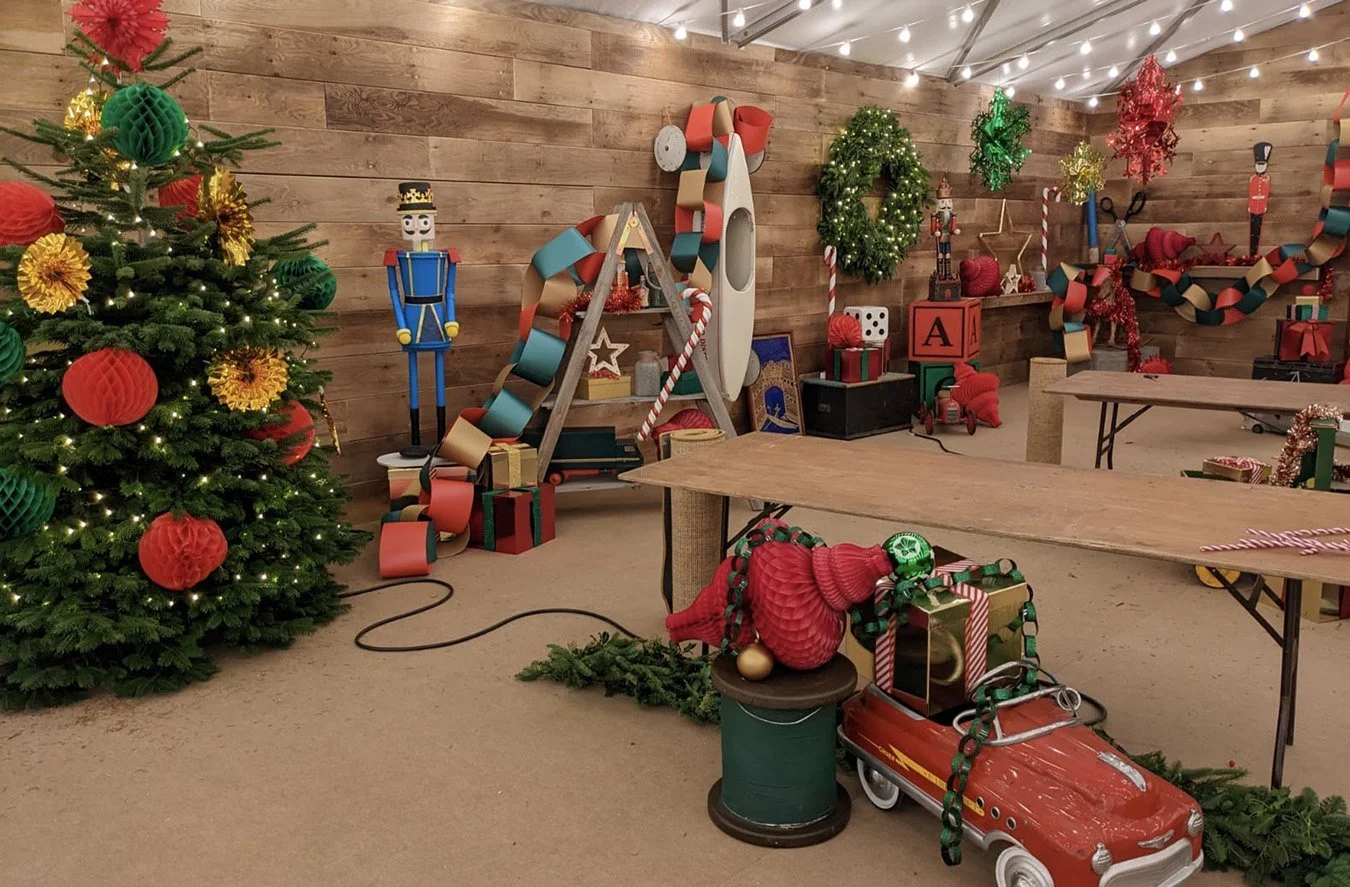 Decorated Christmas party room with Christmas tree, wreath, ornaments, and holiday decorations including a toy car, nutcracker figure, and ribbon garlands.