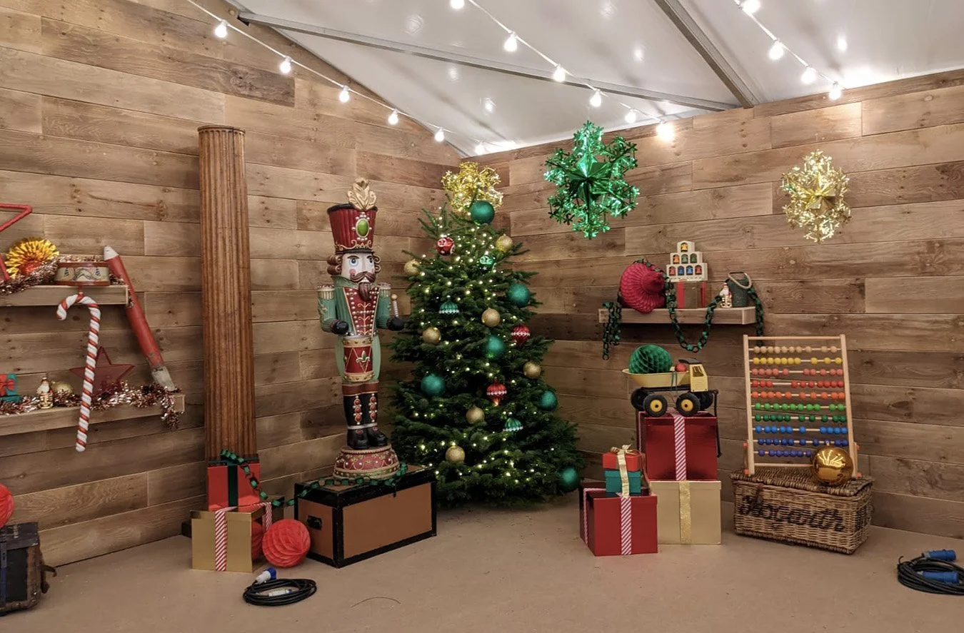 A decorated Christmas scene with a lit Christmas tree, a nutcracker, presents, and holiday decorations on shelves and walls, in a room with wood-paneled walls and string lights overhead.