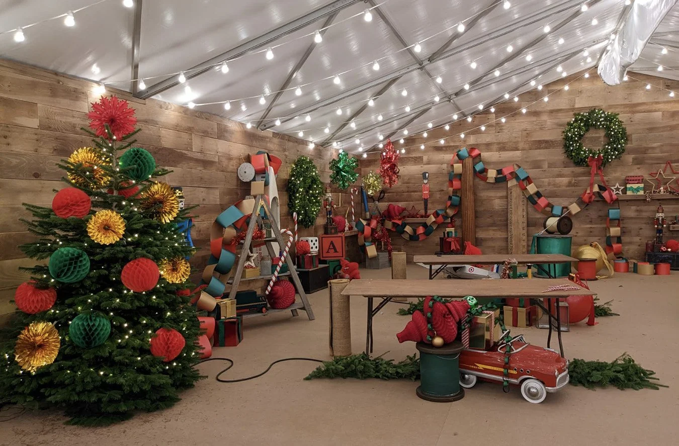 Decorated Christmas tree with red, green, and yellow ornaments, fairy lights, and paper honeycomb balls inside a festive holiday decorated indoor space with wreaths, string lights, and gift boxes.