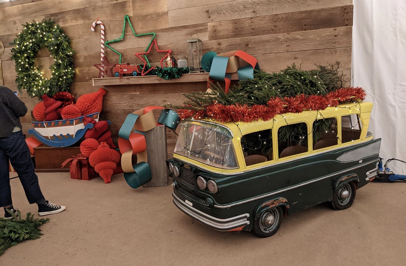 Decorative holiday display with a vintage miniature vehicle decorated with greenery and red tinsel, Christmas wreath, knitted red socks, paper chains, a paper boat, and a wooden wall with star-shaped lights and Christmas ornaments.