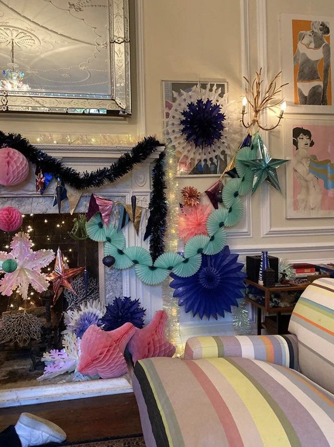 Decorative holiday scene with paper fans in blue, pink, purple, and teal, a black tinsel garland, illuminated string lights, paper lanterns, and artwork on the wall.