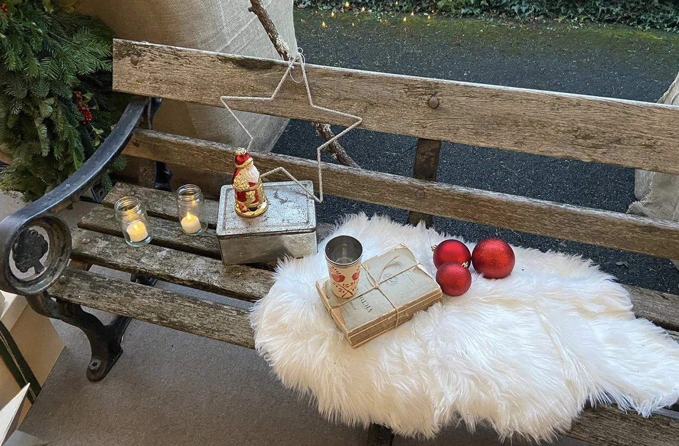 Wooden bench decorated for Christmas with candles, ornaments, books, and a small Santa figurine, with a fluffy white rug on it.