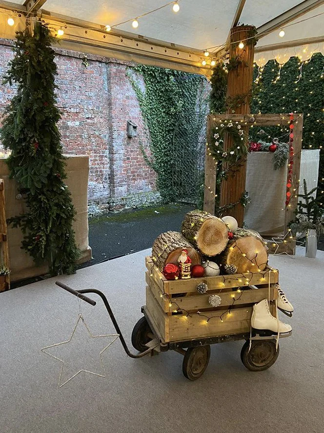 Decorated wooden cart filled with logs, Christmas ornaments, and string lights, surrounded by festive greenery, inside a tent with string lighting and a brick wall outside.