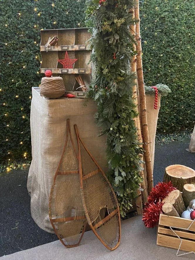 Christmas-themed display with vintage wooden sleds, a cardboard box with greenery and red decorations, a wrapped present, and a rustic setup with fairy lights and greenery.