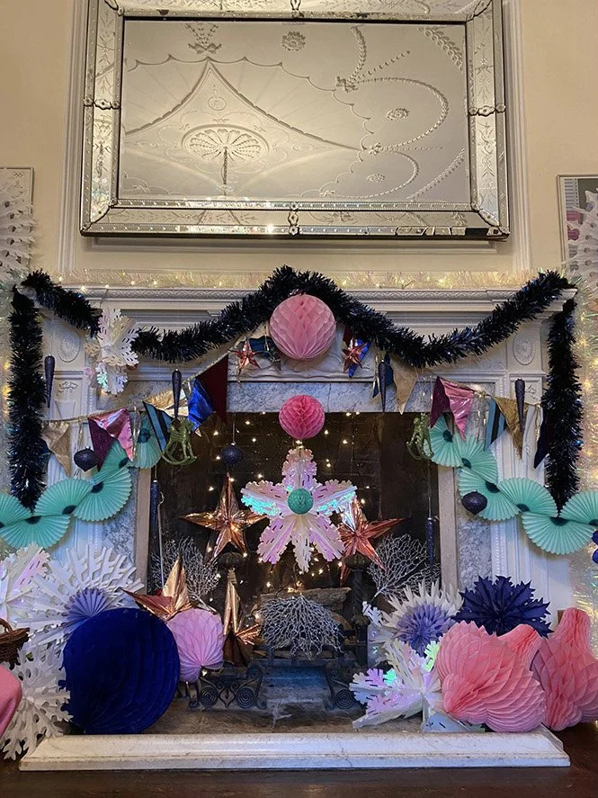 Decorated fireplace with hanging paper lanterns, stars, snowflakes, and paper fans in pastel colors, with a black garland and fairy lights.