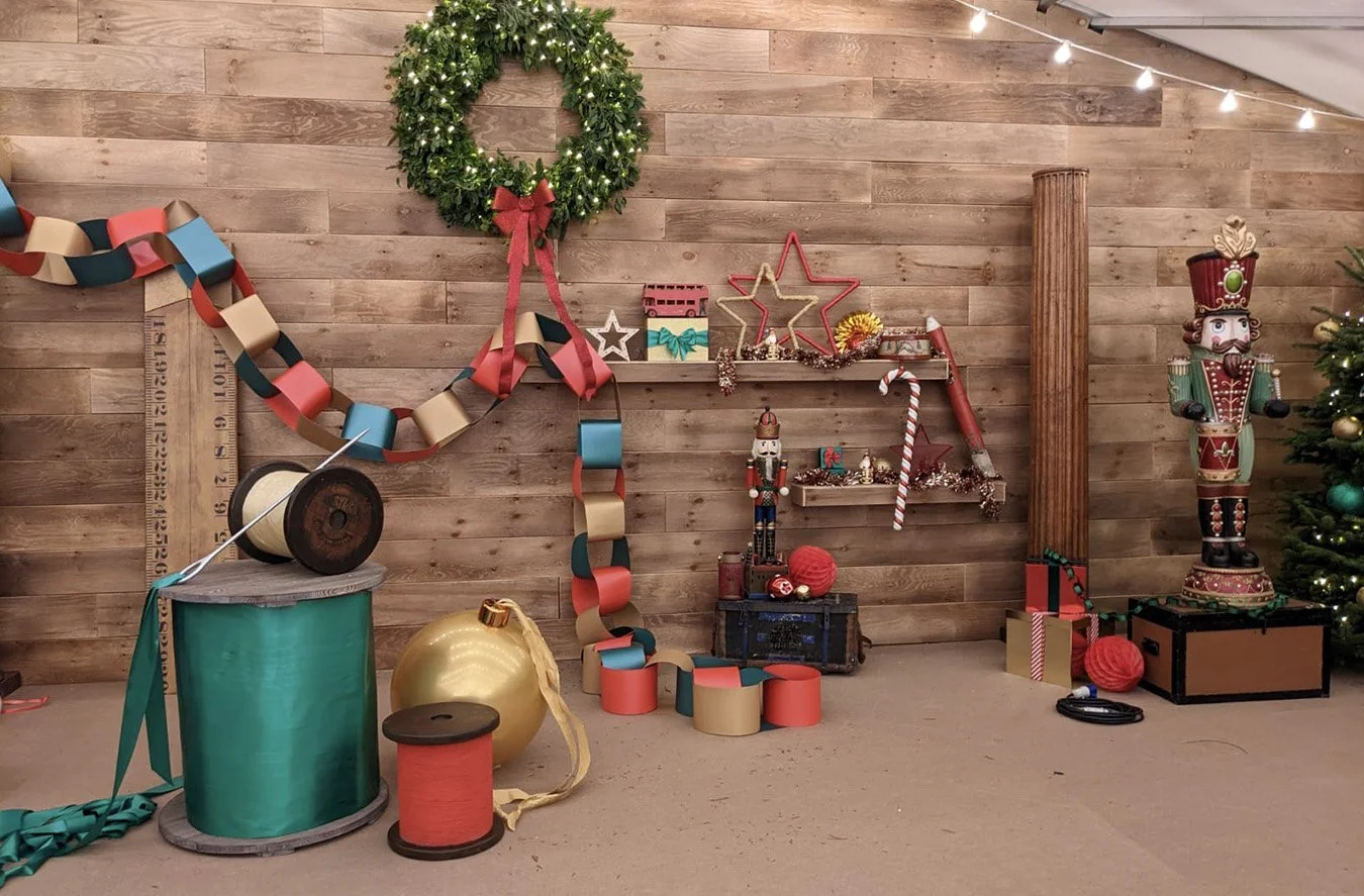 A decorated Christmas scene with a wooden wall background, string lights, a green wreath, paper chain garland, nutcracker figure, Christmas tree with ornaments, and various holiday decorations including stars, gifts, and a red and white candy cane.
