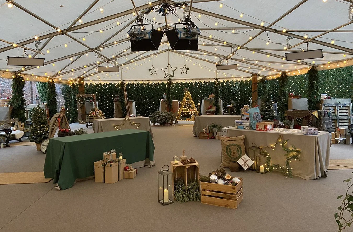 An indoor Christmas market or event space with decorated tables, Christmas trees, string lights, and holiday decorations.