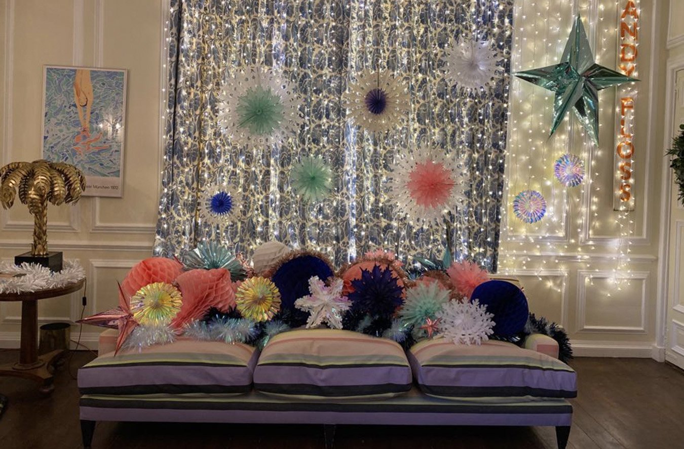 Festive indoor space with a decorated wall featuring string lights, paper flowers, and a large star-shaped decoration. In front, there are colorful paper lanterns, tinsel, and a striped couch.