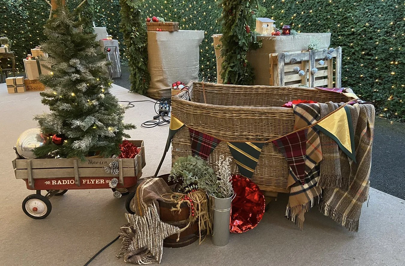 Decorative Christmas scene with a small Christmas tree in a wagon, festive garland, plaid scarves, ornaments, wrapped gifts, and string lights in the background.