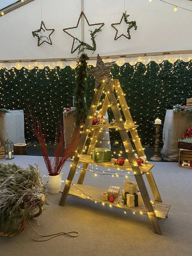 Christmas-themed display with a wooden ladder decorated with warm string lights and ornaments, a large star on top, surrounded by wrapped gifts, festive faux foliage, and star-shaped decorations hanging from the ceiling. Background features a dark, s