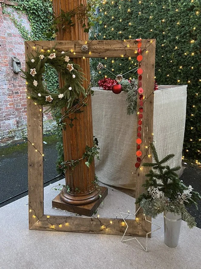 A wooden picture frame decorated with holiday ornaments, string lights, and greenery stands around a wooden column. There are Christmas ornaments, a wreath, and a small potted evergreen tree with frosted pinecones, placed in a silver pot, in front of