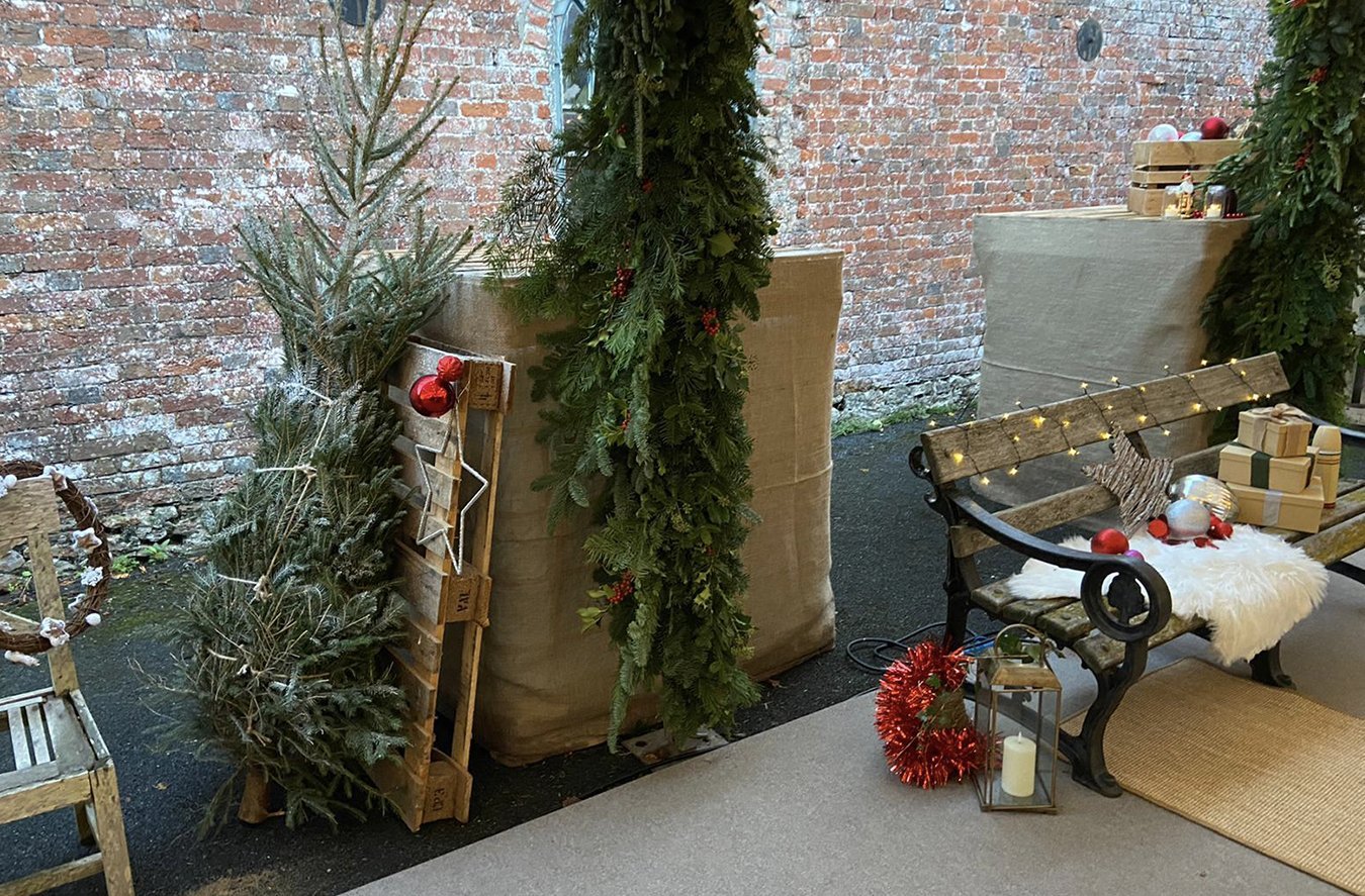 Decorated outdoor Christmas display with greenery, ornaments, presents, and holiday lights, featuring a bench with festive decorations, a lantern, and a brick wall in the background.