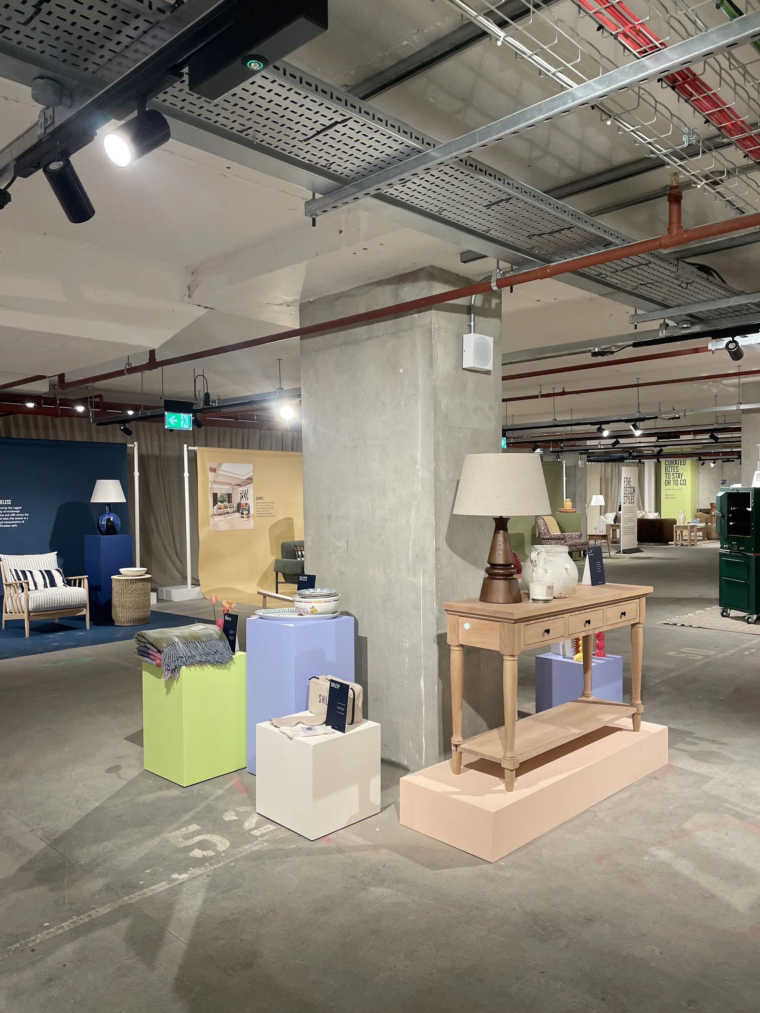 Display of various home furniture and decor items in a showroom, including a wooden console table with a lamp, vases, and decorative objects, plus chairs, lamps, and artwork in the background.