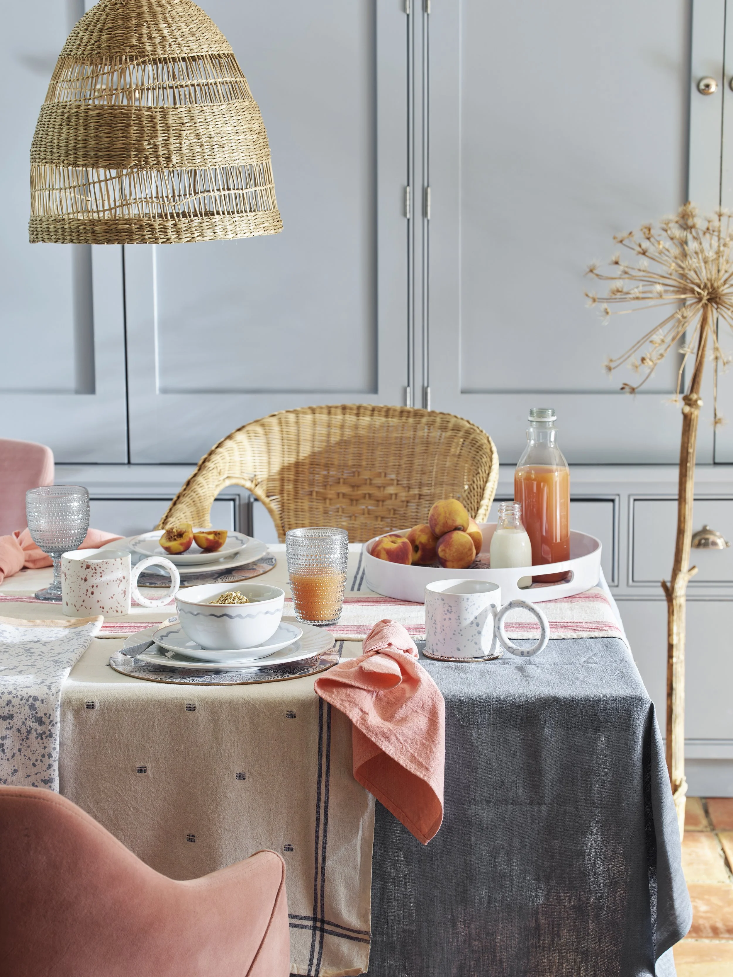 A cozy breakfast table set with cups, bowls, plates, and a variety of fruits including peaches. The table has a layered tablecloth with different patterns and colors, and pink napkins. In the background, there are blue cabinets, and a woven pendant l