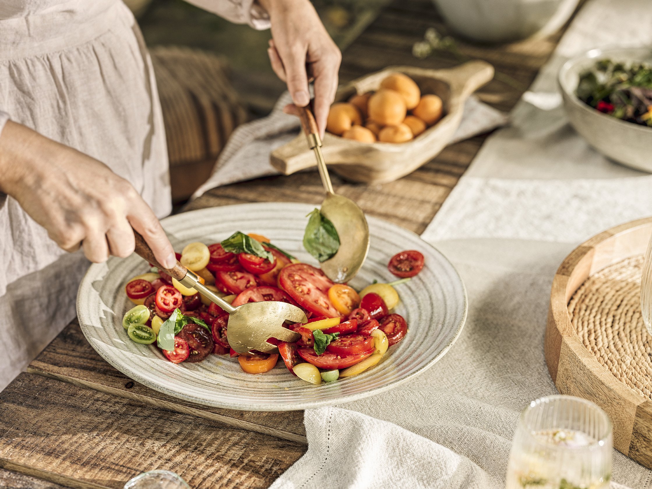 A person serving a fresh tomato salad onto a plate at a rustic outdoor table, with bowls of salad ingredients in the background.