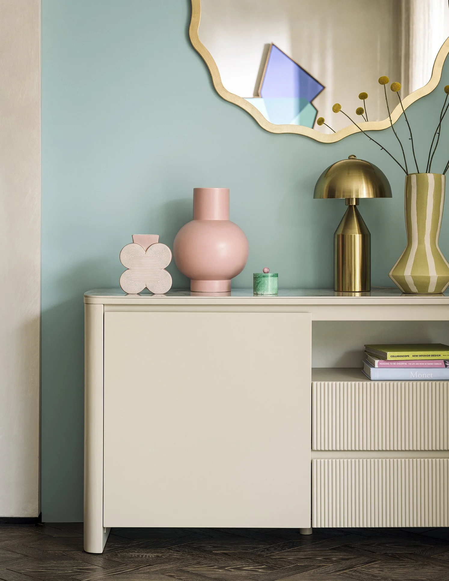 Decorative objects on a white sideboard against a blue-green wall, including pink vases, a brass lamp, a striped vase with yellow flowers, and a shelf with books, with a wavy mirror above.