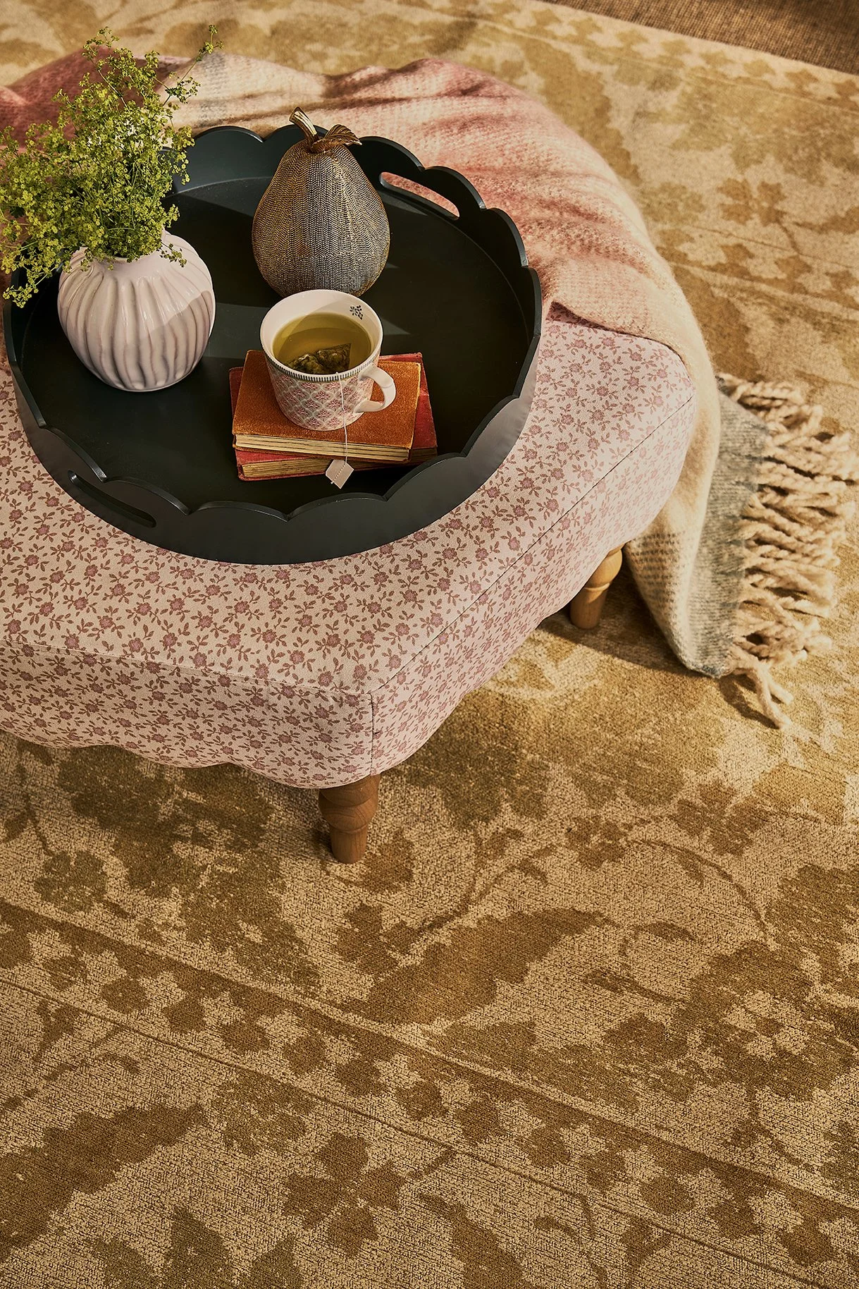 Tray with a potted plant, a decorative pumpkin, and a cup of tea on a pink patterned ottoman with a blanket and floral rug.
