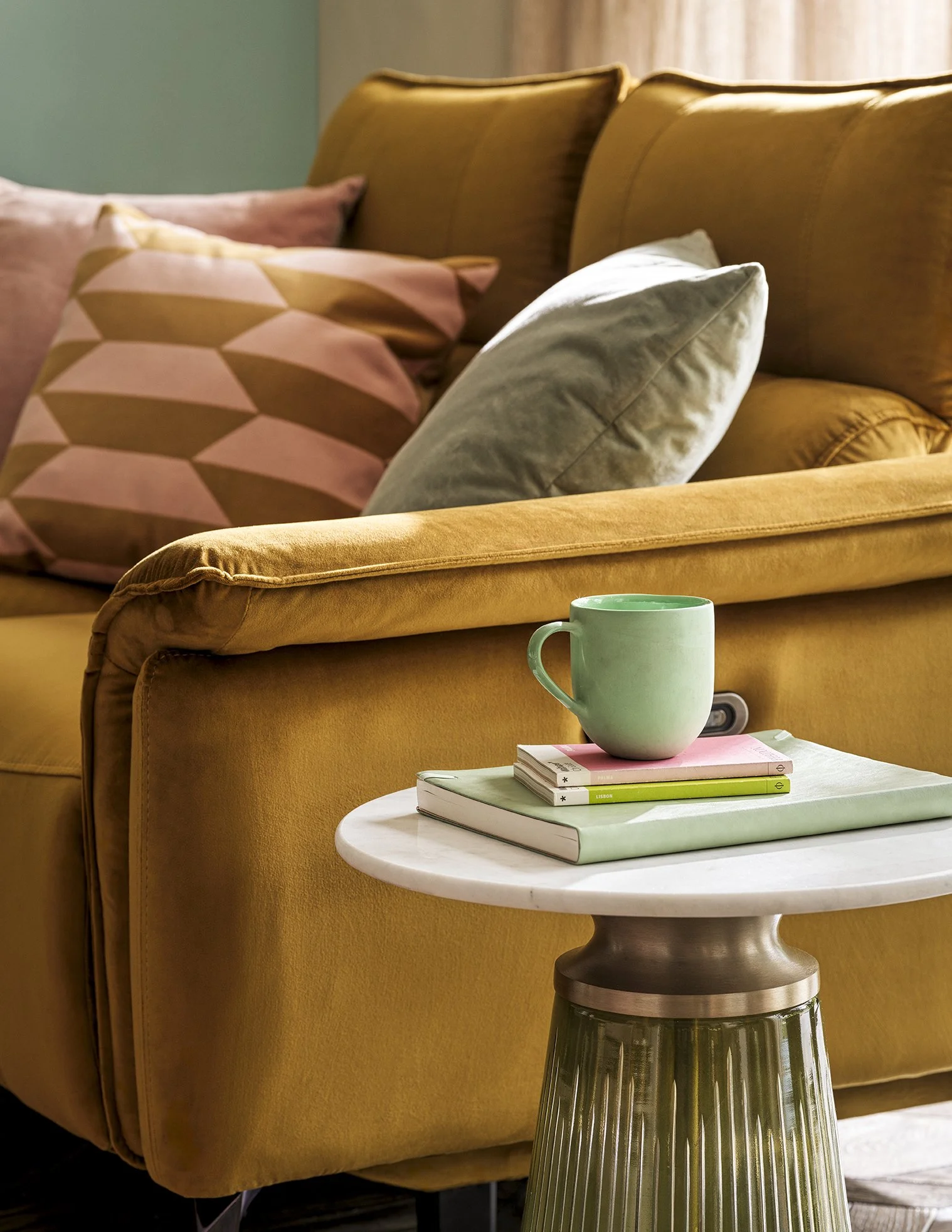 A cozy living room scene with a mustard yellow sofa with two cushions, one pink with geometric pattern and one plain grey, and a small white marble side table holding a green mug and a few books.