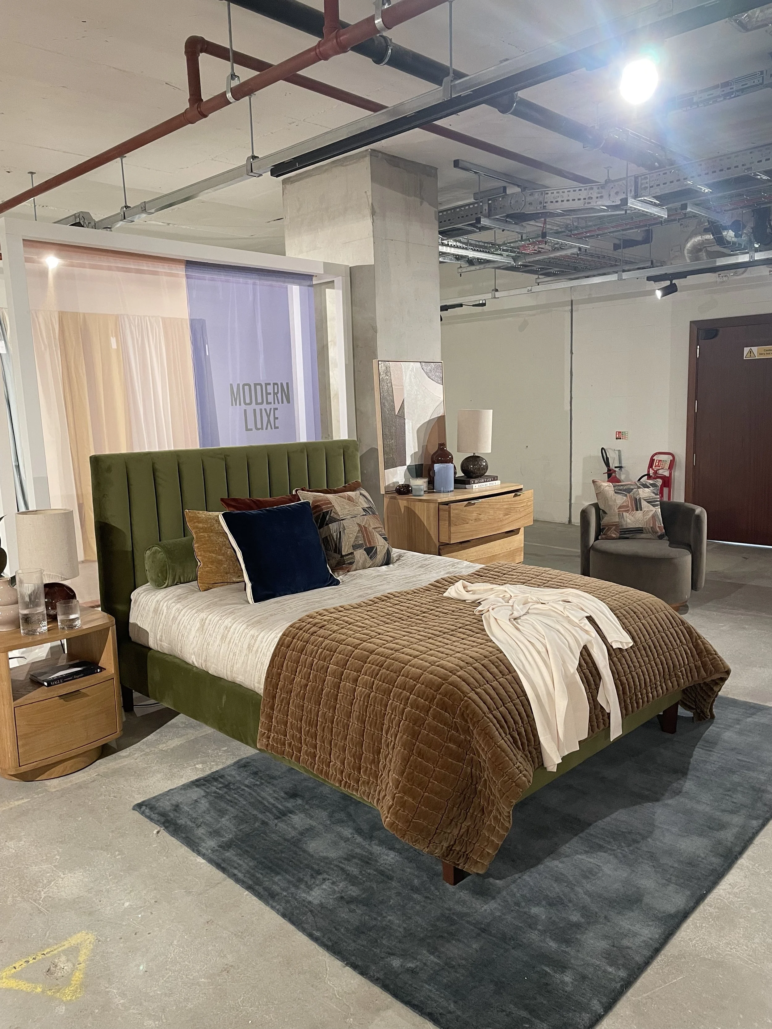 A bedroom setup showcases a green upholstered bed with a beige and brown quilt, various patterned pillows, and a white sheet on top. There are wooden nightstands with lamps on either side, and a wooden dresser with decorative vases and a lamp behind 