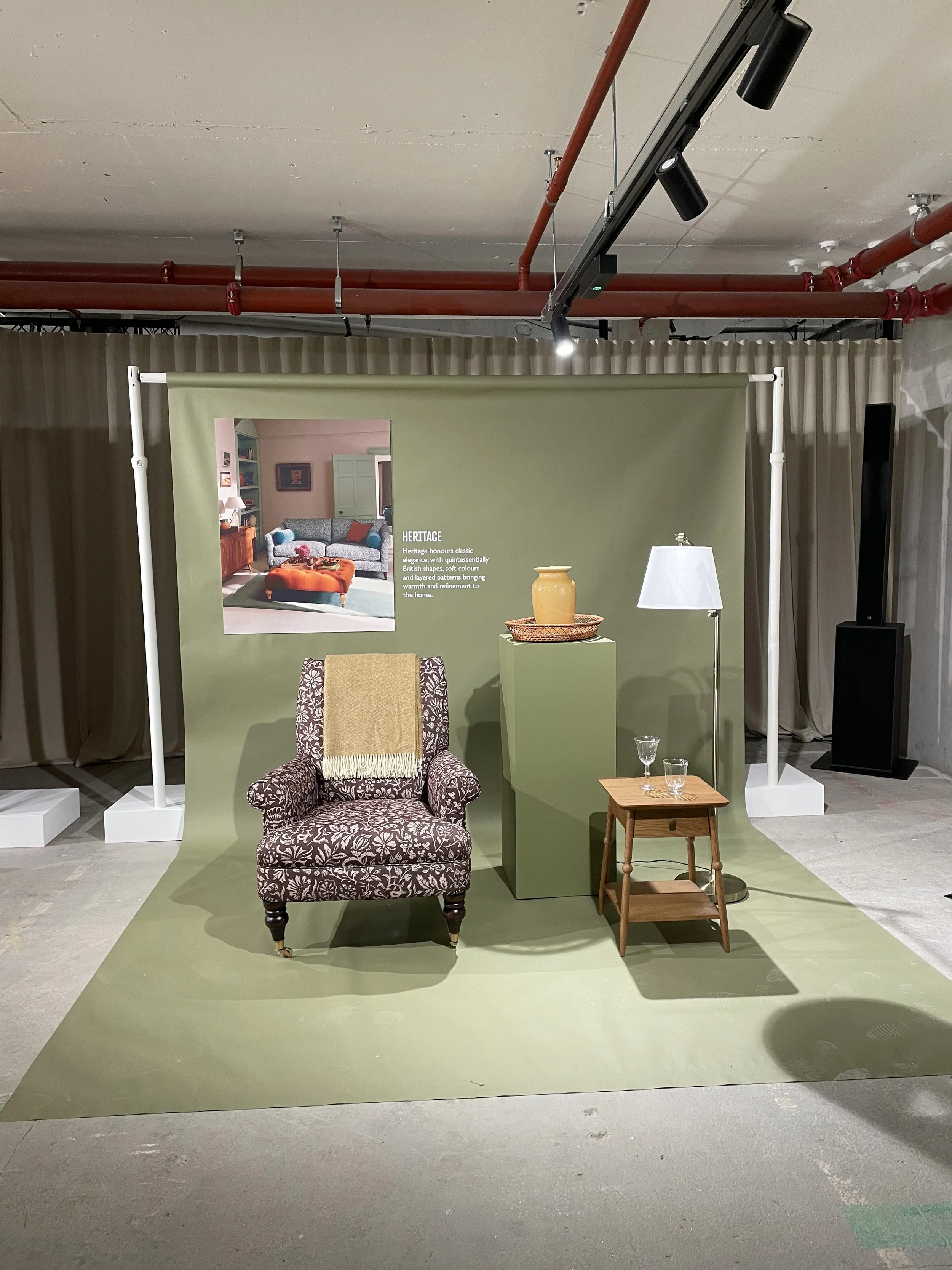 Interior scene featuring a vintage armchair with a tan throw blanket, a small wooden side table with two glasses, a floor lamp, and a green display stand with a ceramic pitcher on top, set against a green backdrop with a furniture advertisement poste