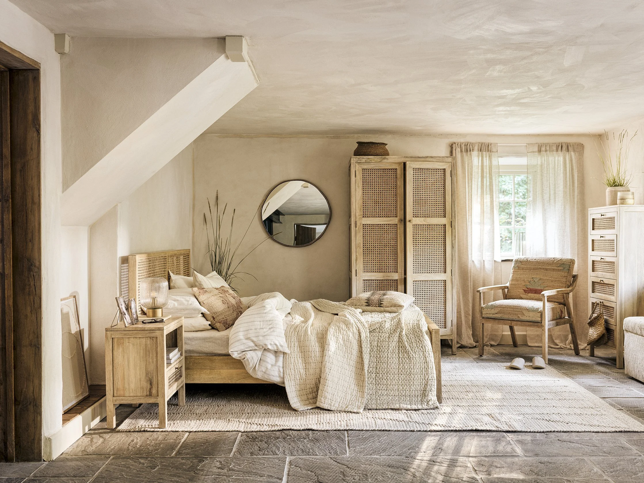 A cozy bedroom with a bed, nightstand, chair, dresser, and a window with sheer curtains, decorated in neutral tones and natural light.